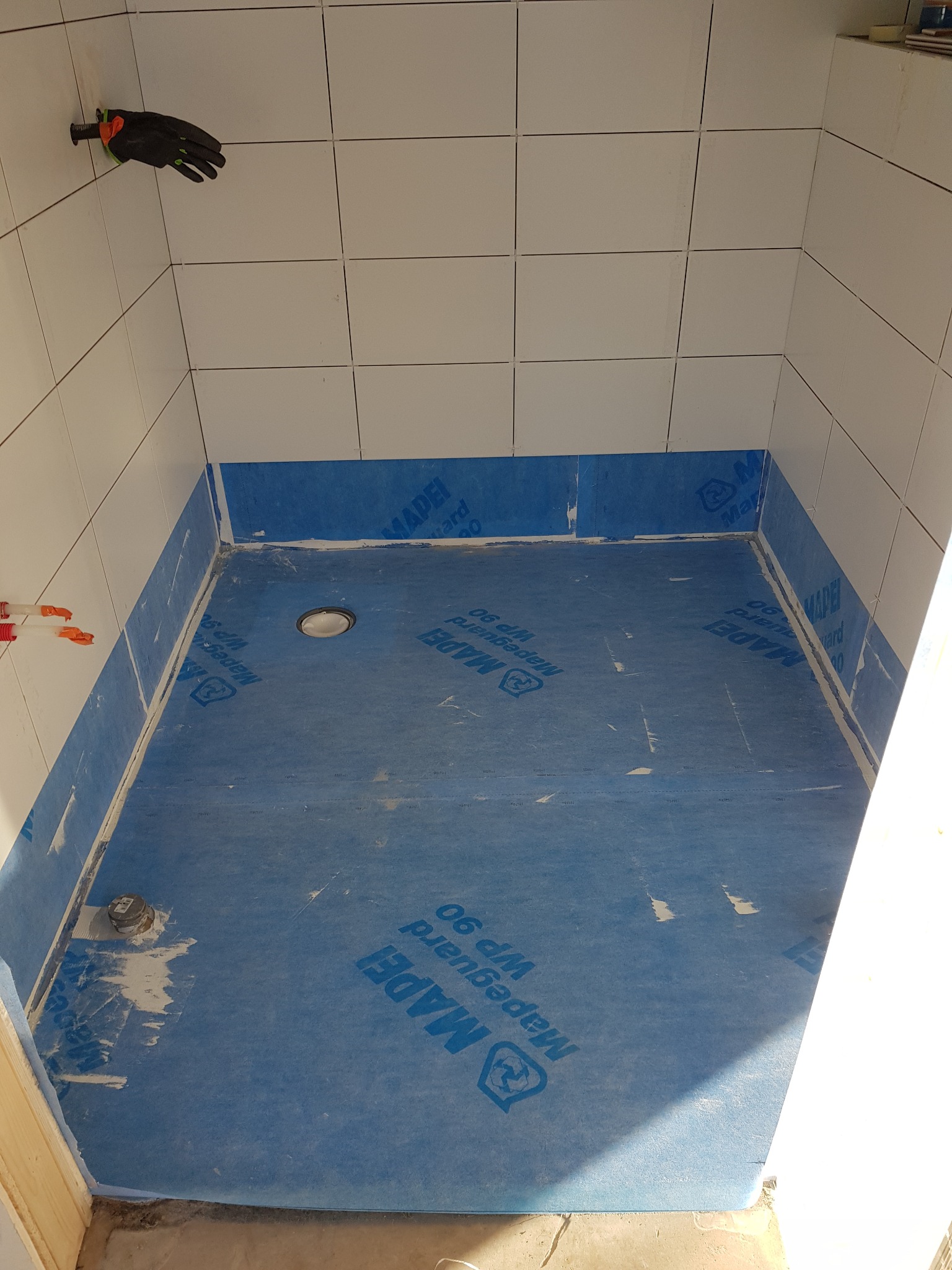 Przygotowanie brodzika: hydroizolacja z folii w płynie Mapeguard WP 90 na podłodze i ścianach, białe płytki na ścianach, rękawica robocza zawieszona na ścianie.