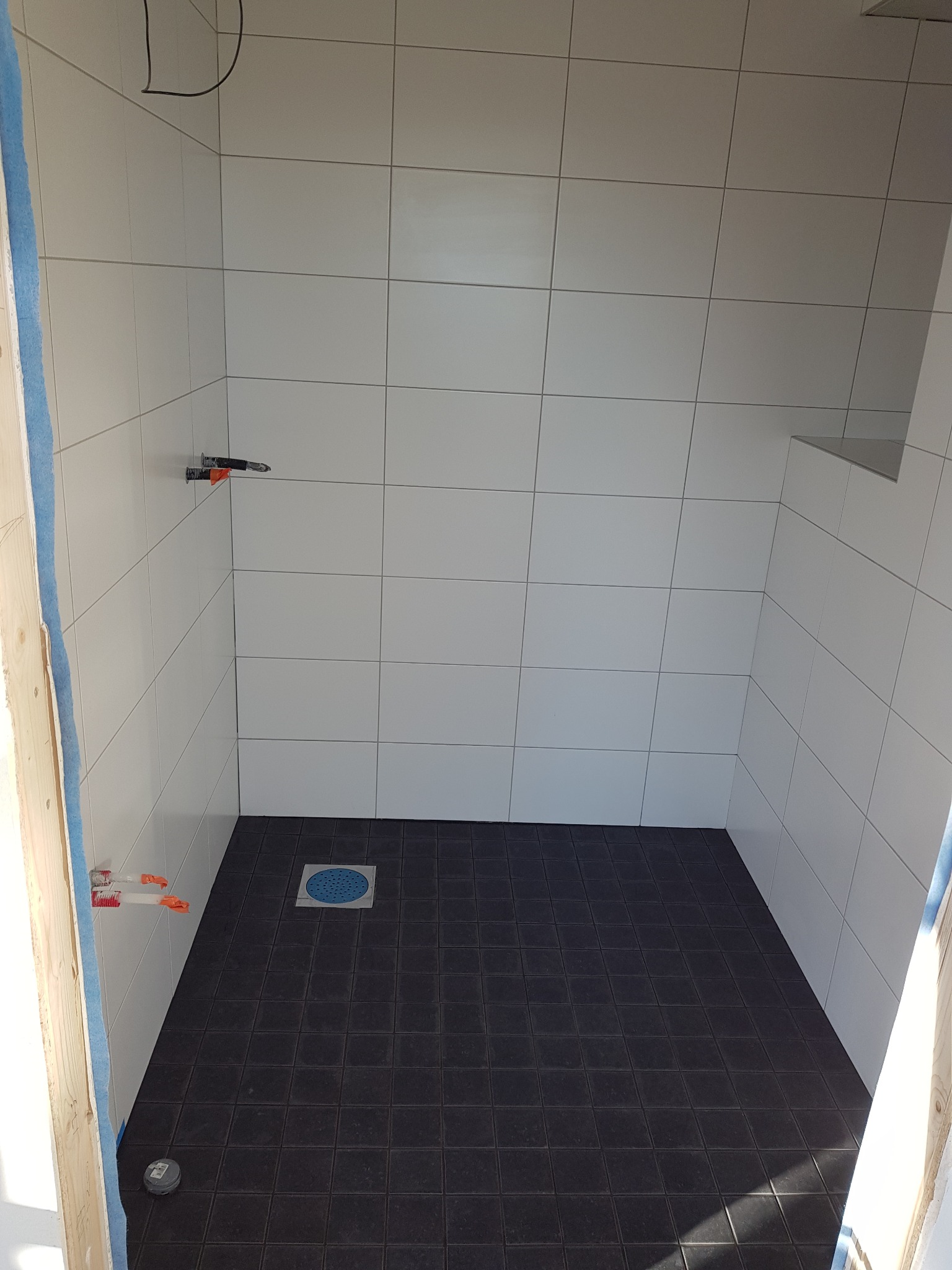 Niedokończona kabina prysznicowa z białymi płytkami ściennymi i ciemną podłogą z odpływem, widoczne wystające rury instalacji wodnej z zamontowanymi zaciskami do płytek.