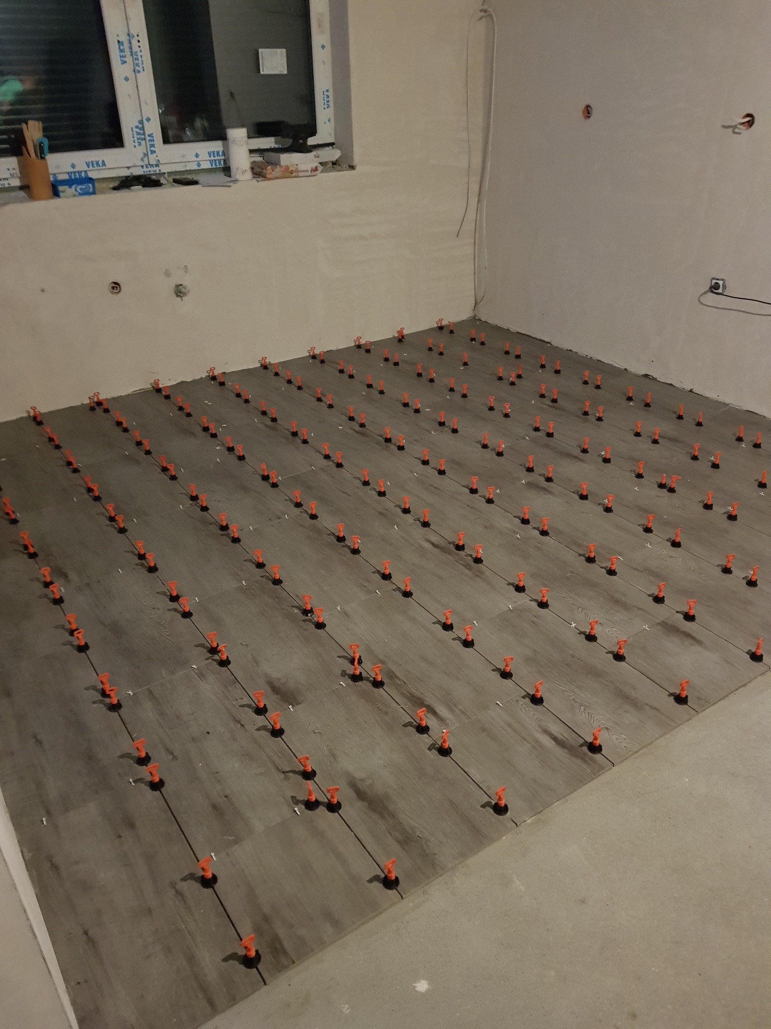 Układanie płytek podłogowych w jasnym pomieszczeniu, z użyciem pomarańczowych klipsów poziomujących, widoczne niedokończone ściany i okno.