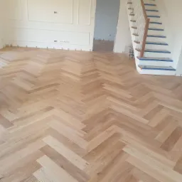 Ułożony parkiet w jodełkę francuską z jasnego drewna w przestronnym wnętrzu z białymi ścianami i schodami zabezpieczonymi taśmą malarską.