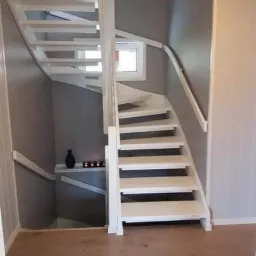 Białe, drewniane schody kręcone w szarym wnętrzu, z widoczną półką z dekoracjami i małym oknem w tle.