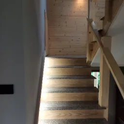 Jasne, drewniane schody z szarymi nakładkami dywanowymi, oświetlone pojedynczym reflektorem na drewnianej ścianie w tle.