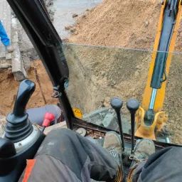 Widok z kabiny operatora koparki podczas prac ziemnych: wykop, sterta piasku, pracownik w czerwonych rękawicach przy wykopie.