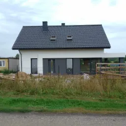 Nowoczesny dom jednorodzinny z szarą elewacją i dachem z dachówki, widoczne okna dachowe, teren wokół domu częściowo zagospodarowany.