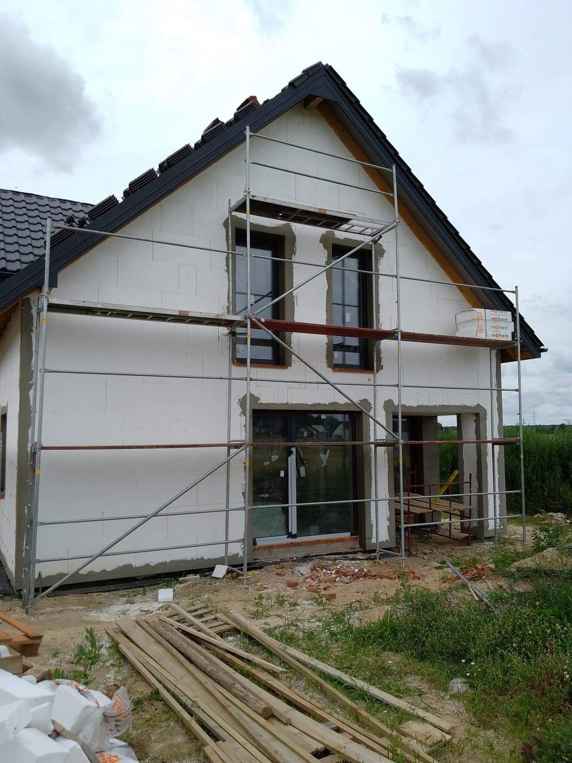 Dom jednorodzinny w trakcie ocieplania, widoczne rusztowania i białe płyty izolacyjne na elewacji, czarna dachówka. Częściowo ocieplona ściana szczytowa.