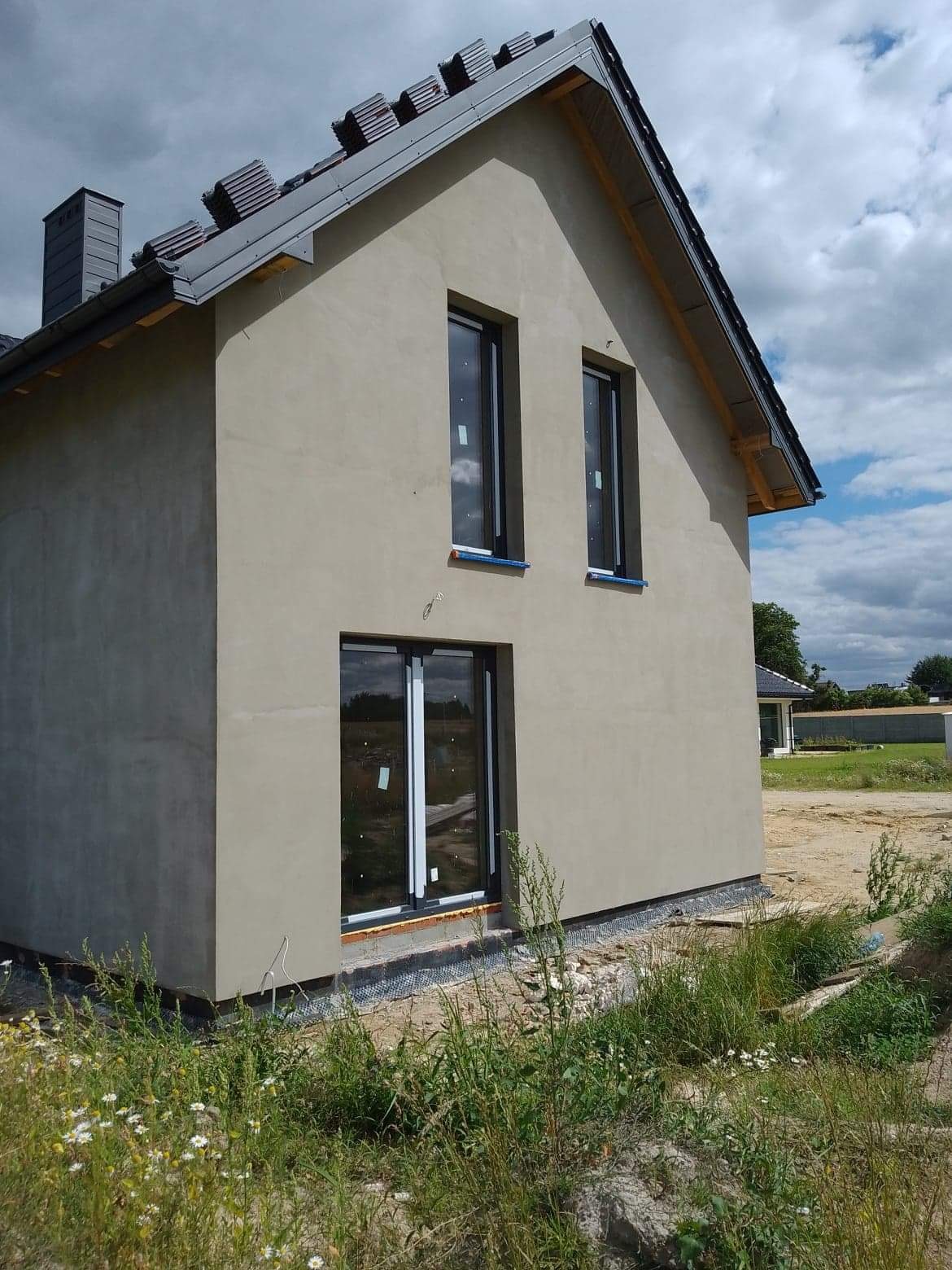 Surowa elewacja domu jednorodzinnego w trakcie ocieplania, widoczne czarne okna i przygotowane podłoże pod tynk, zarys dachu z dachówkami, w tle zachmurzone niebo.