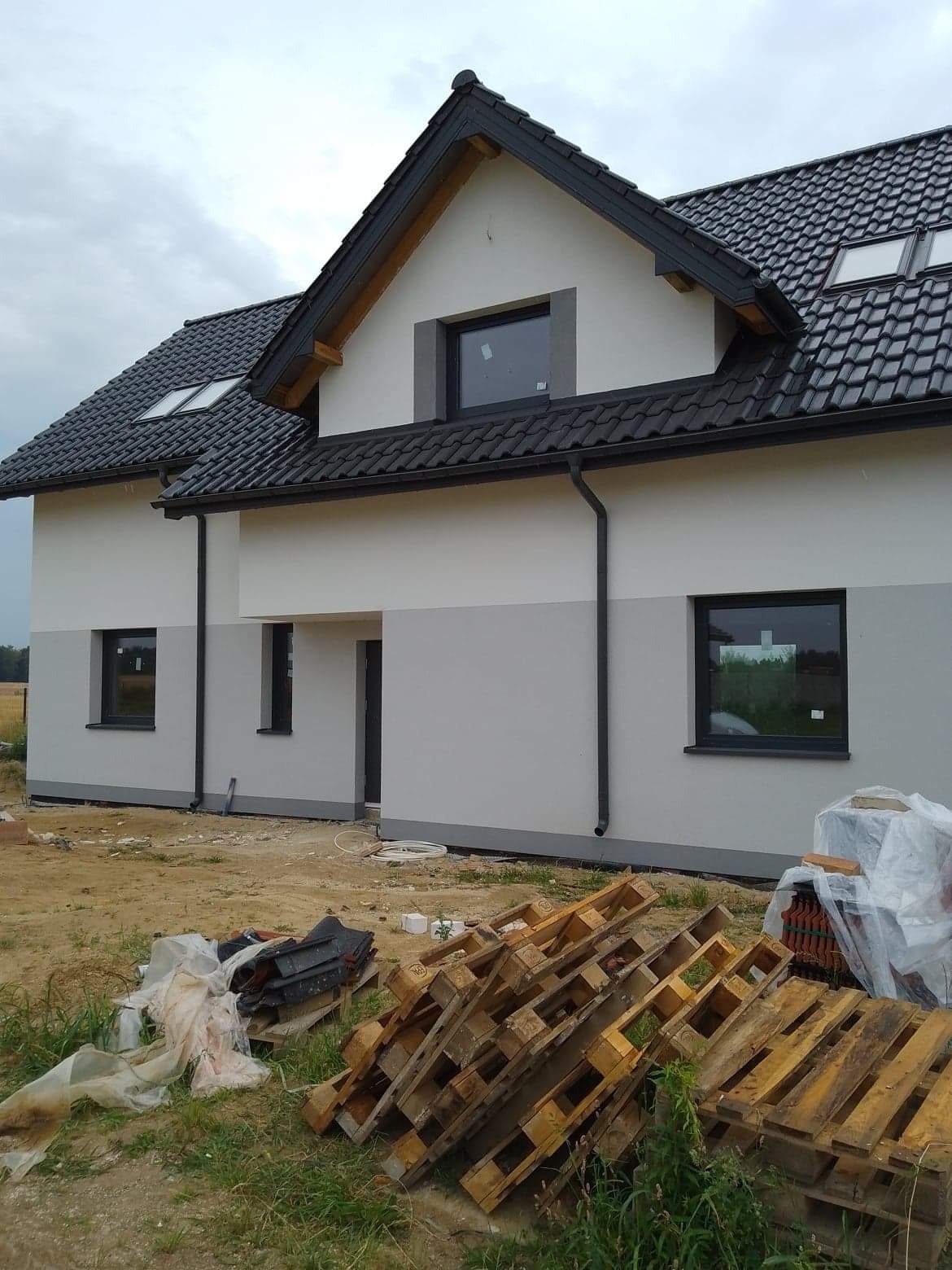 Dwukondygnacyjny dom jednorodzinny z ocieploną elewacją w odcieniach szarości i bieli, czarna dachówka, widoczne palety i materiały budowlane na trawiastym podłożu.