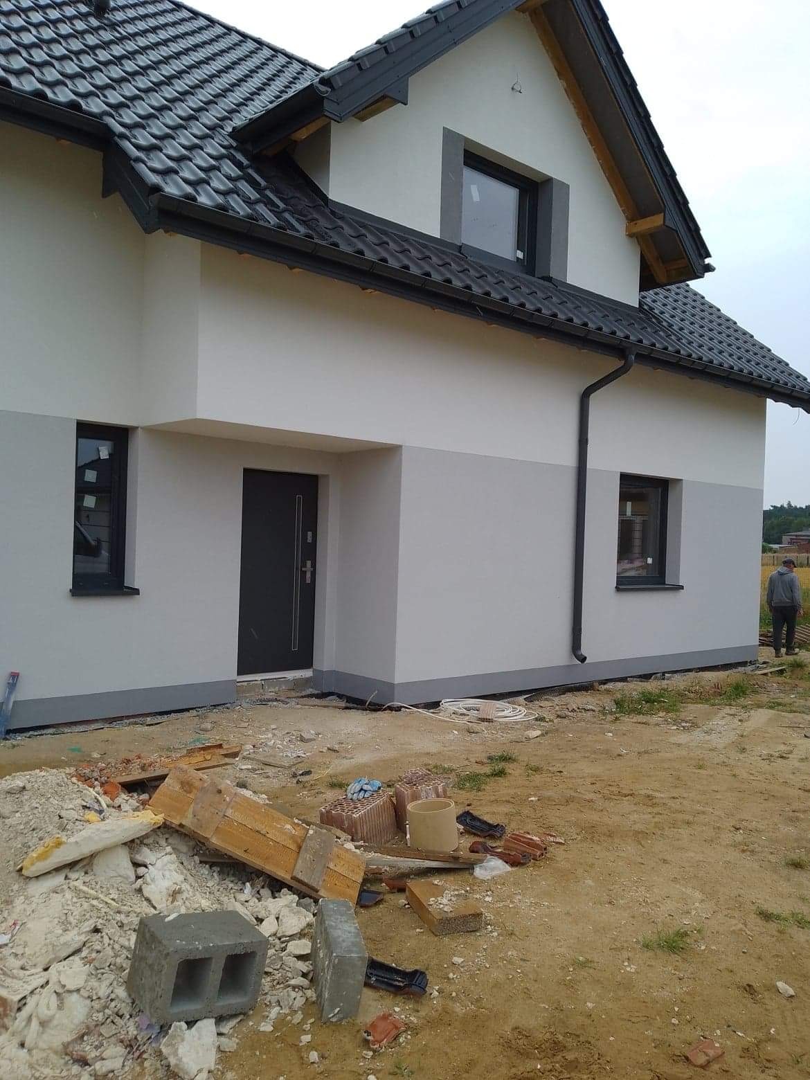 Dwukolorowa elewacja domu jednorodzinnego z ciemną stolarką okienną i drzwiami, widoczne gruz i materiały budowlane na posesji.