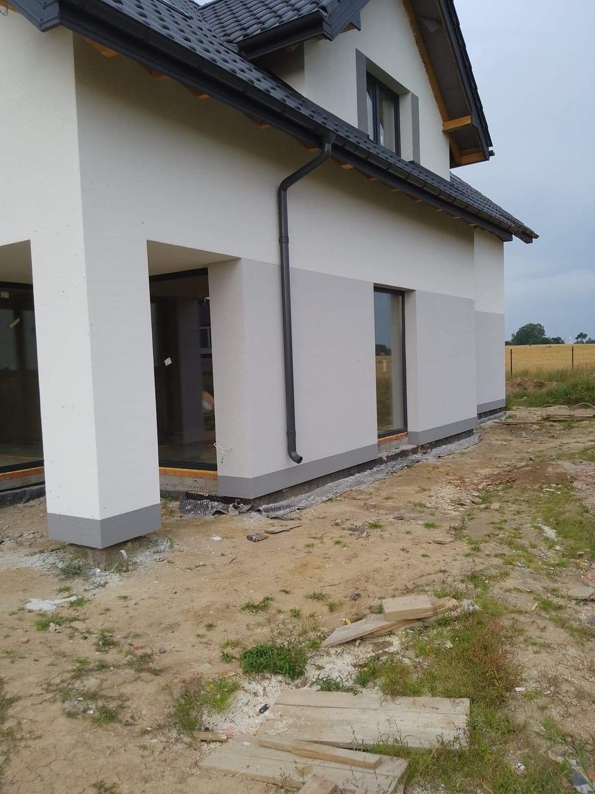 Dwukolorowa elewacja domu jednorodzinnego z szarą podmurówką i grafitową rynną, widok z boku na tle pola.