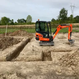 Pomarańczowa mini koparka Kubota KX019-4 na placu budowy, wykonująca wykop pod fundamenty, widoczne wykopane rowy, w tle drzewa i słup wysokiego napięcia.