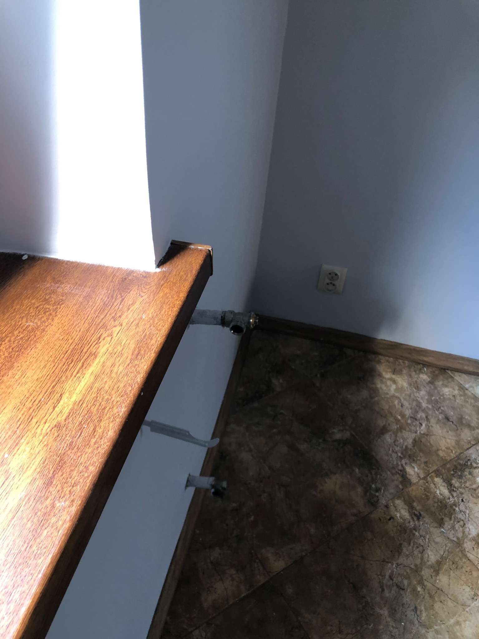 Niedokończone wykończenie: drewniany blat, instalacja hydrauliczna wystająca ze ściany, marmurowe płytki podłogowe i gniazdko elektryczne na niebieskiej ścianie.