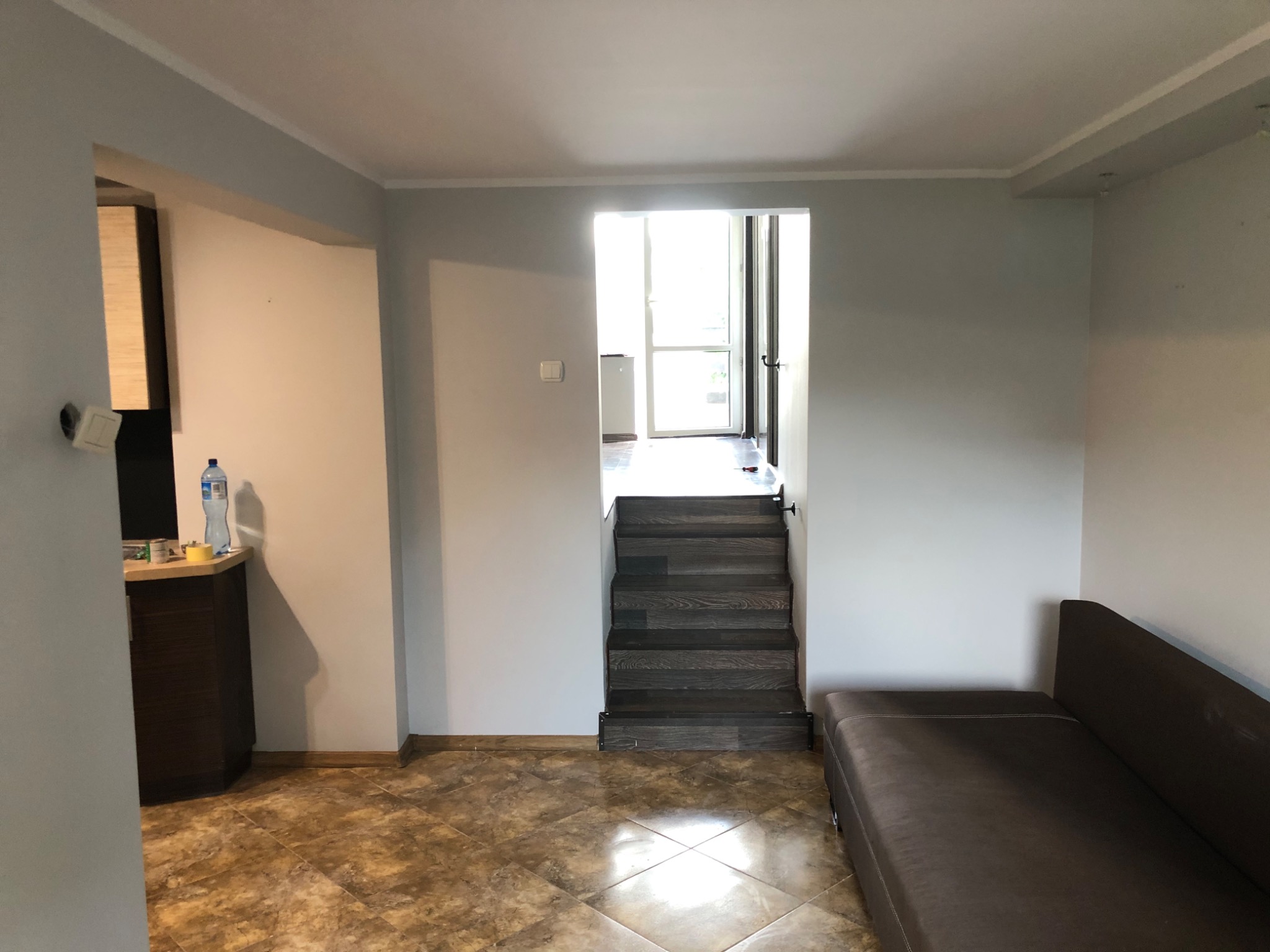 Wnętrze pokoju z brązową sofą, schodami prowadzącymi do przeszklonych drzwi balkonowych i brązową podłogą w romby, z fragmentem zabudowy kuchennej po lewej stronie.