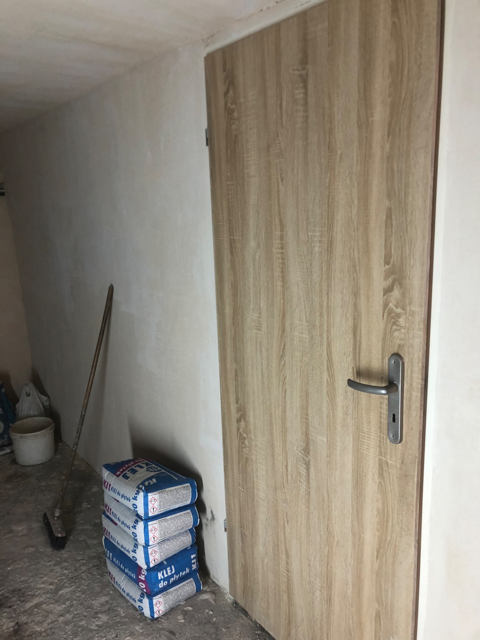 Wnętrze w trakcie remontu: drzwi z okleiną drewnopodobną, ściana po tynkowaniu, worki z klejem do płytek i miotła.