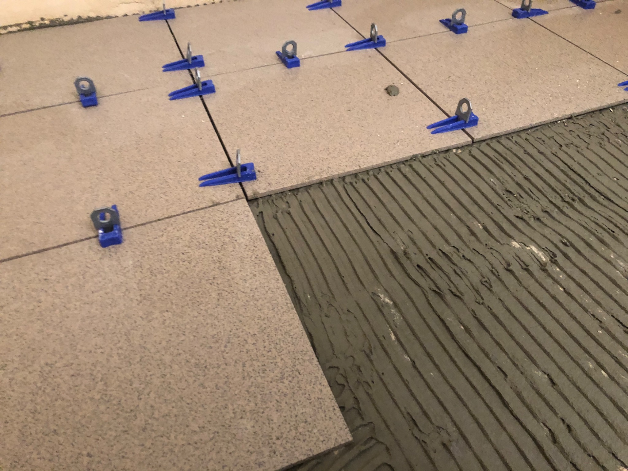 Układanie płytek podłogowych z użyciem systemu poziomowania: widoczna zaprawa klejowa w rowkach i niebieskie klipsy stabilizujące.