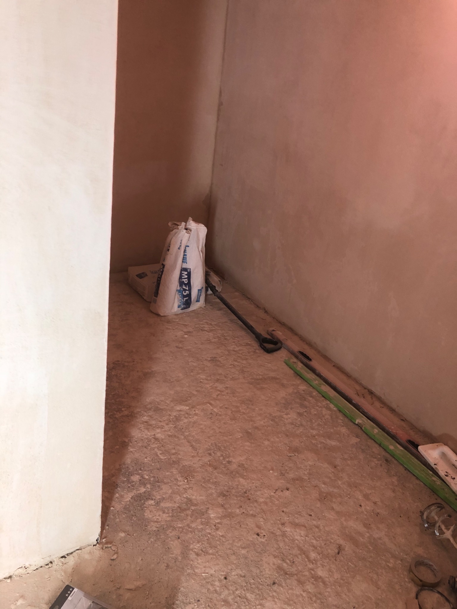 Wnętrze w trakcie tynkowania: ściany pokryte świeżym tynkiem, worek z zaprawą MP 75, poziomica i mieszadło leżące na podłodze.