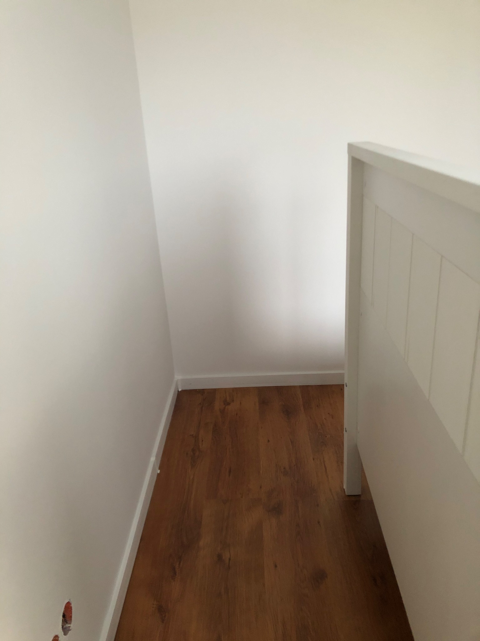 Narożnik pomieszczenia z drewnianą podłogą, białe ściany z listwami przypodłogowymi i widocznym otworem po gniazdku elektrycznym.