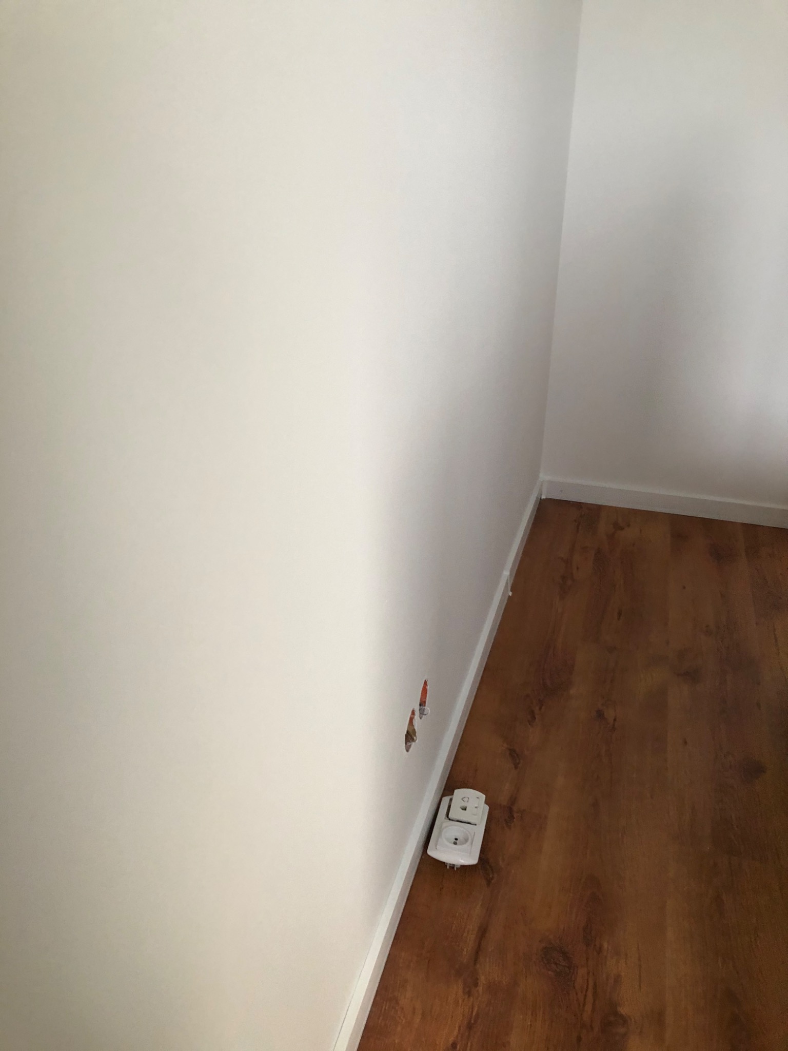 Narożnik pokoju po malowaniu ścian na biało, widoczne gniazdka elektryczne i podłoga z paneli imitujących drewno, z widocznymi niedoskonałościami w postaci otworów w ścianie.