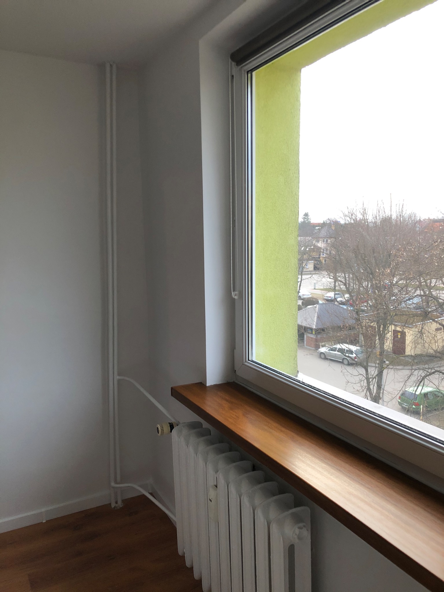 Wnętrze pokoju z białymi ścianami, drewnianą podłogą i nowym oknem z zielonym obramowaniem, pod którym zamontowano drewniany parapet i biały grzejnik.