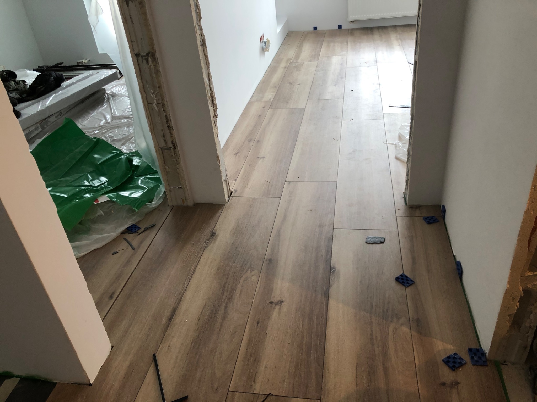 Układanie podłogi z płytek imitujących drewno w korytarzu, widoczne plastikowe krzyżyki dystansowe, w tle folia ochronna i materiały budowlane.