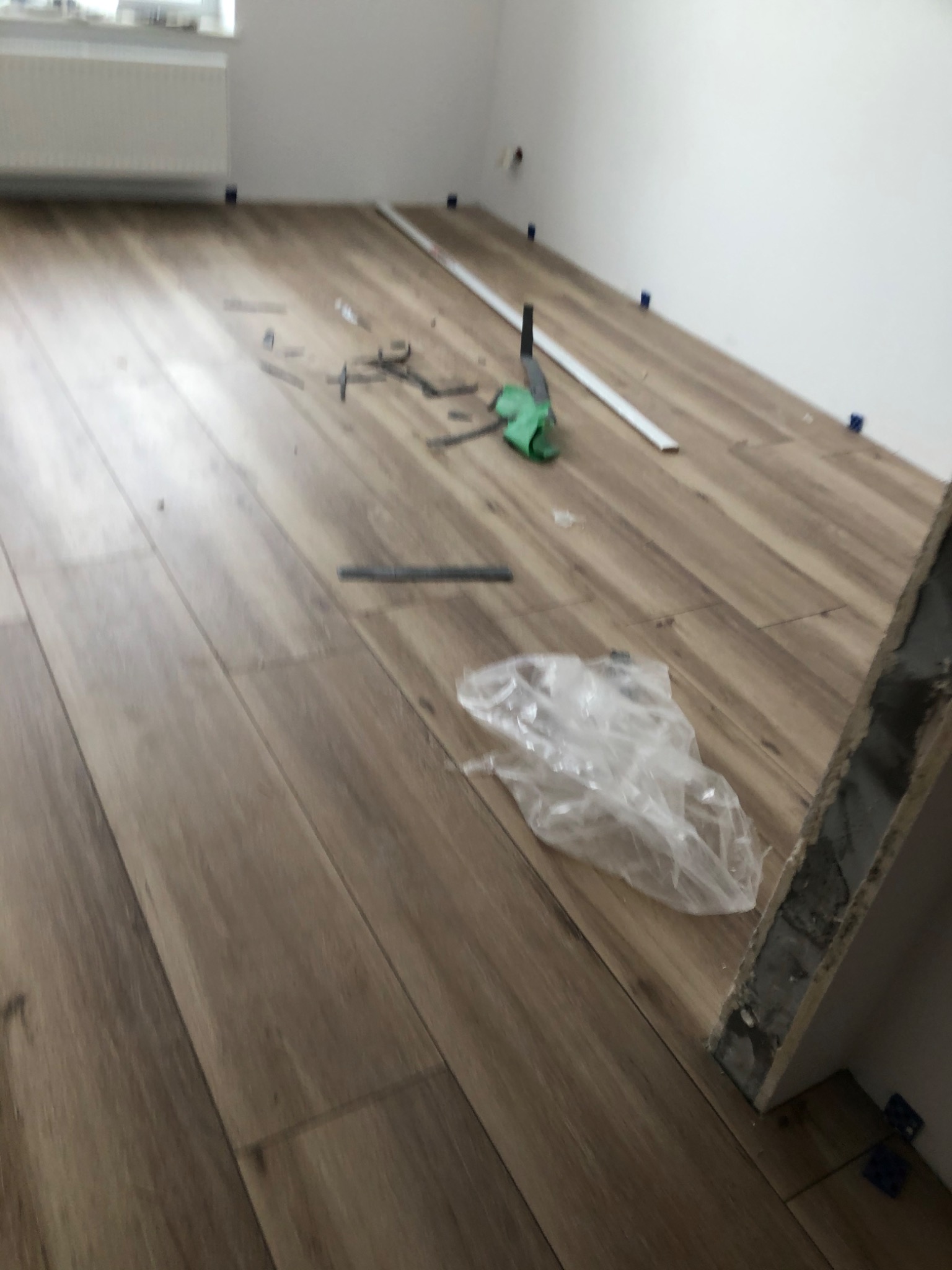 Układanie paneli podłogowych imitujących drewno, widoczne narzędzia glazurnicze, listwa przypodłogowa, niebieskie kliny dystansowe i worek folowy na podłodze w trakcie montażu.