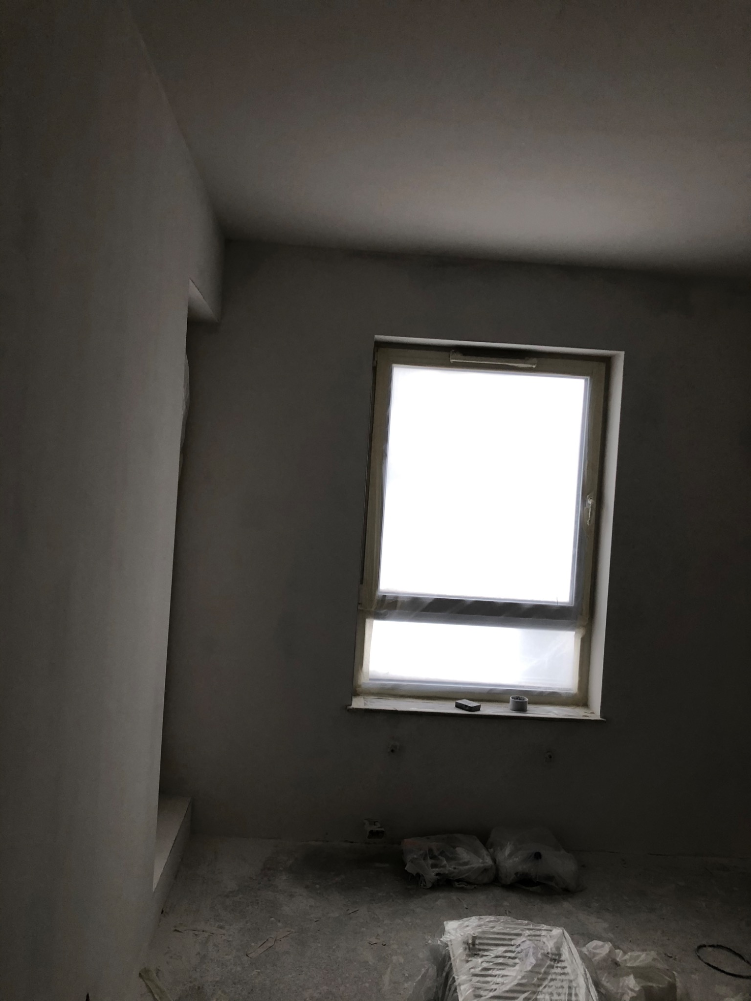 Pokój w trakcie prac wykończeniowych: świeżo otynkowane ściany, okno zabezpieczone folią, widoczne materiały budowlane na podłodze.