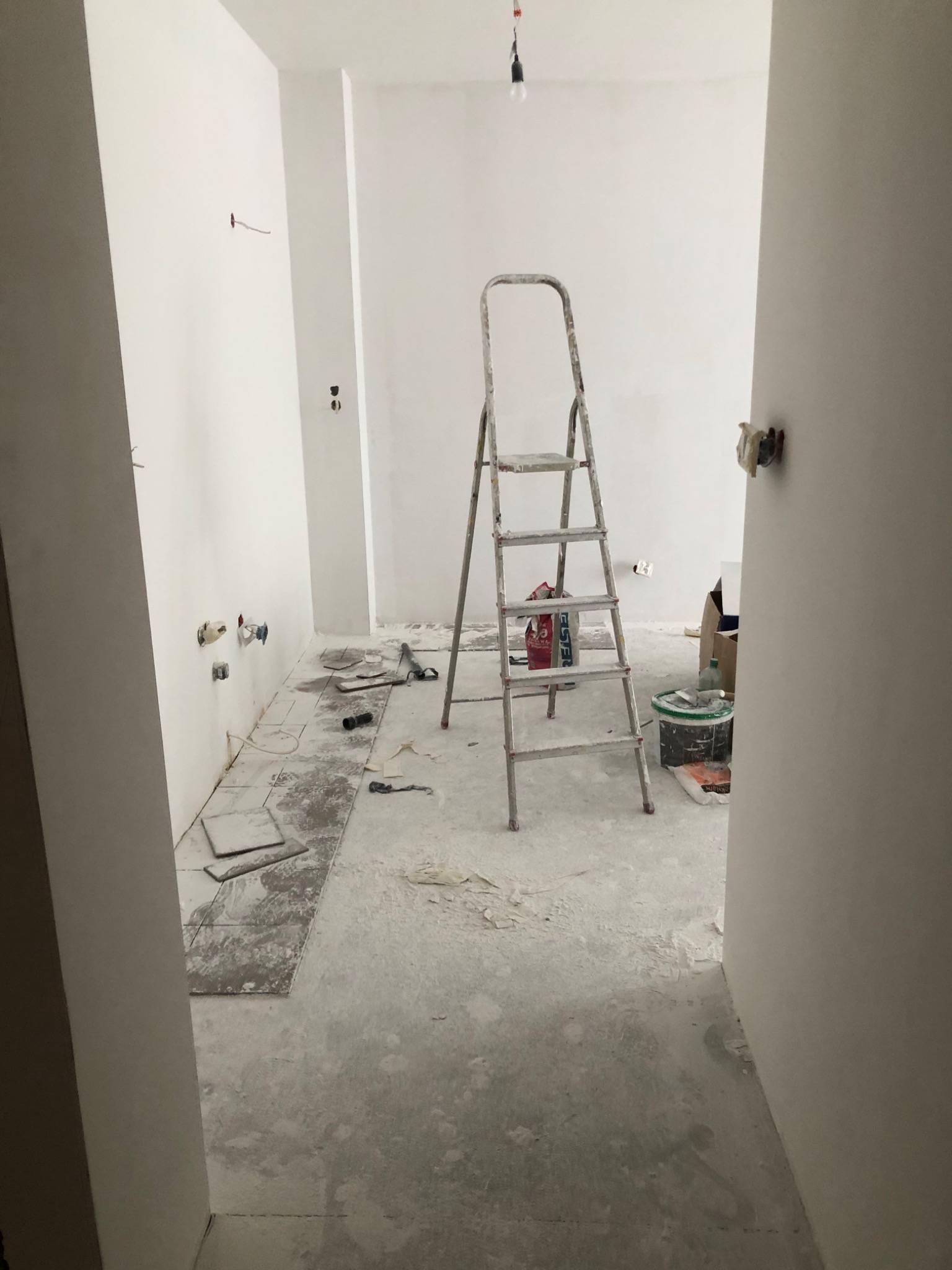 Pokój w trakcie remontu: białe ściany, surowy beton na podłodze, drabina malarska, widoczne instalacje elektryczne i hydrauliczne, płytki podłogowe przygotowane do ułożenia.