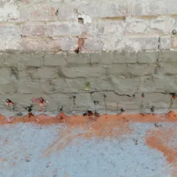 Ceglany fundament z nałożoną warstwą szarego betonu wzmocnioną metalowymi kotwami, widoczne ślady pomarańczowej farby na betonie poniżej.