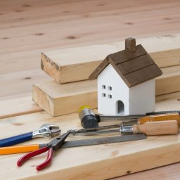 FOXX Sp. zo.o. - Drewniane deski, model domu i narzędzia: kombinerki z czerwonymi uchwytami, klucz nastawny z niebieskim uchwytem, pilnik, śrubokręty z drewnianymi uchwytami i gumowy młotek na jasnym drewnianym tle.