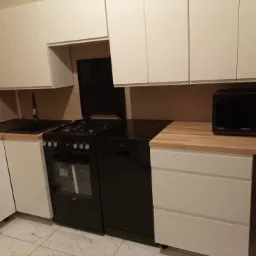 Nowoczesna kuchnia z białymi szafkami, czarnym zlewem, kuchenką i mikrofalówką, blat drewnopodobny, marmurowa podłoga.