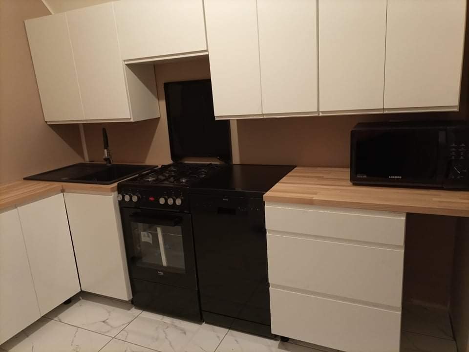 Nowoczesna kuchnia z białymi szafkami, czarnym zlewem, kuchenką i mikrofalówką, blat drewnopodobny, marmurowa podłoga.