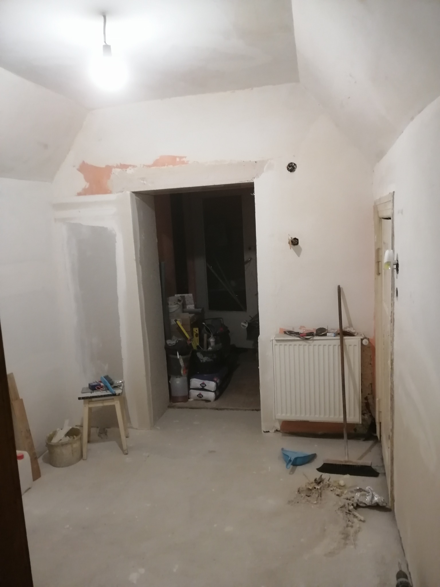 Pokój w trakcie remontu: ściany częściowo pokryte gładzią, widoczne ślady po wcześniejszych pracach, narzędzia i materiały budowlane w tle, surowe oświetlenie.