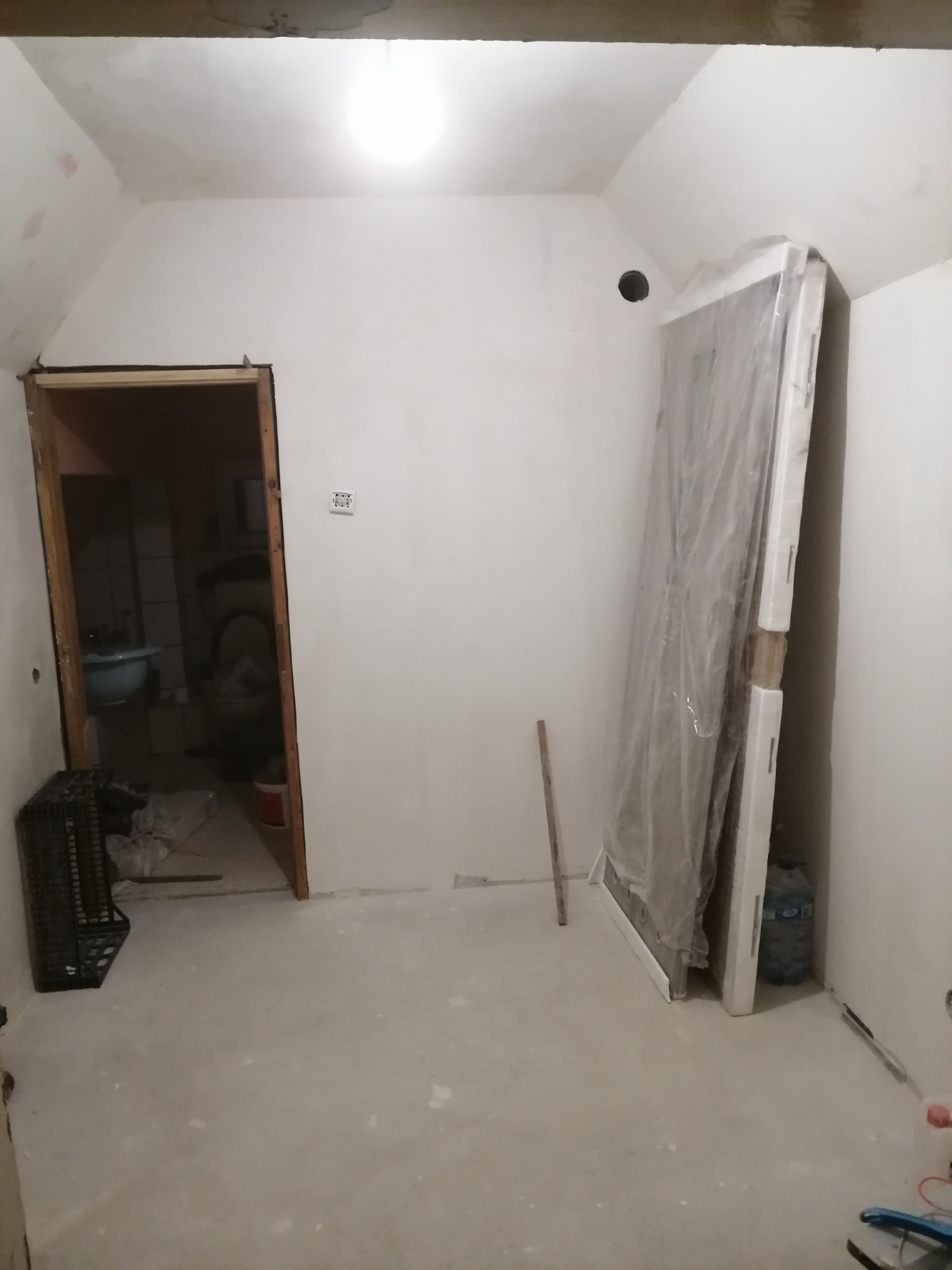 Przygotowane pomieszczenie do prac wykończeniowych: surowe ściany, w rogu oparta o ścianę nowa ościeżnica drzwiowa zabezpieczona folią, widoczna łazienka w tle.