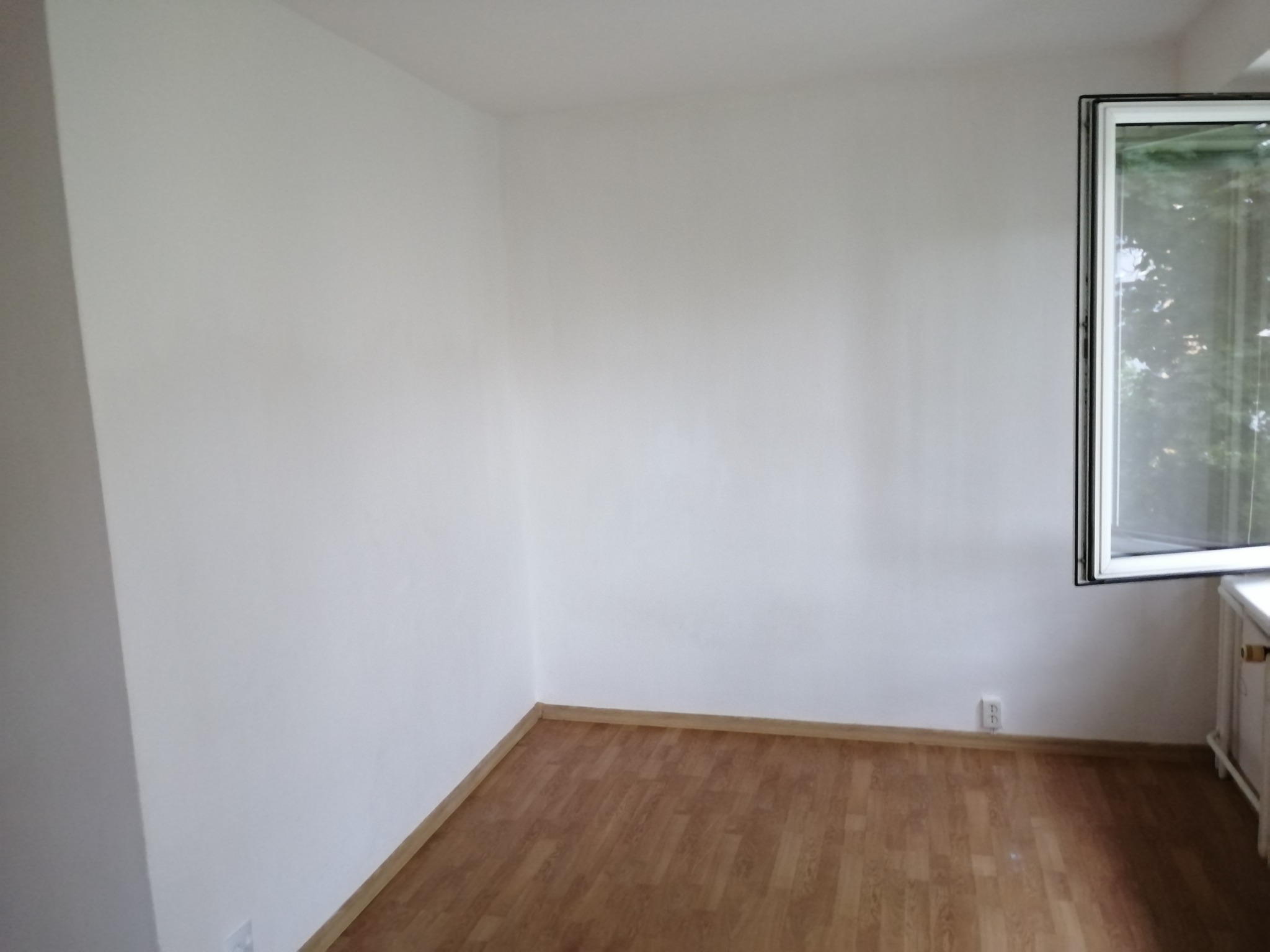 Pusty pokój po remoncie: świeżo pomalowane białe ściany, jasnobrązowa podłoga imitująca drewno, otwarte okno z widokiem na zieleń na zewnątrz, widoczny fragment grzejnika.