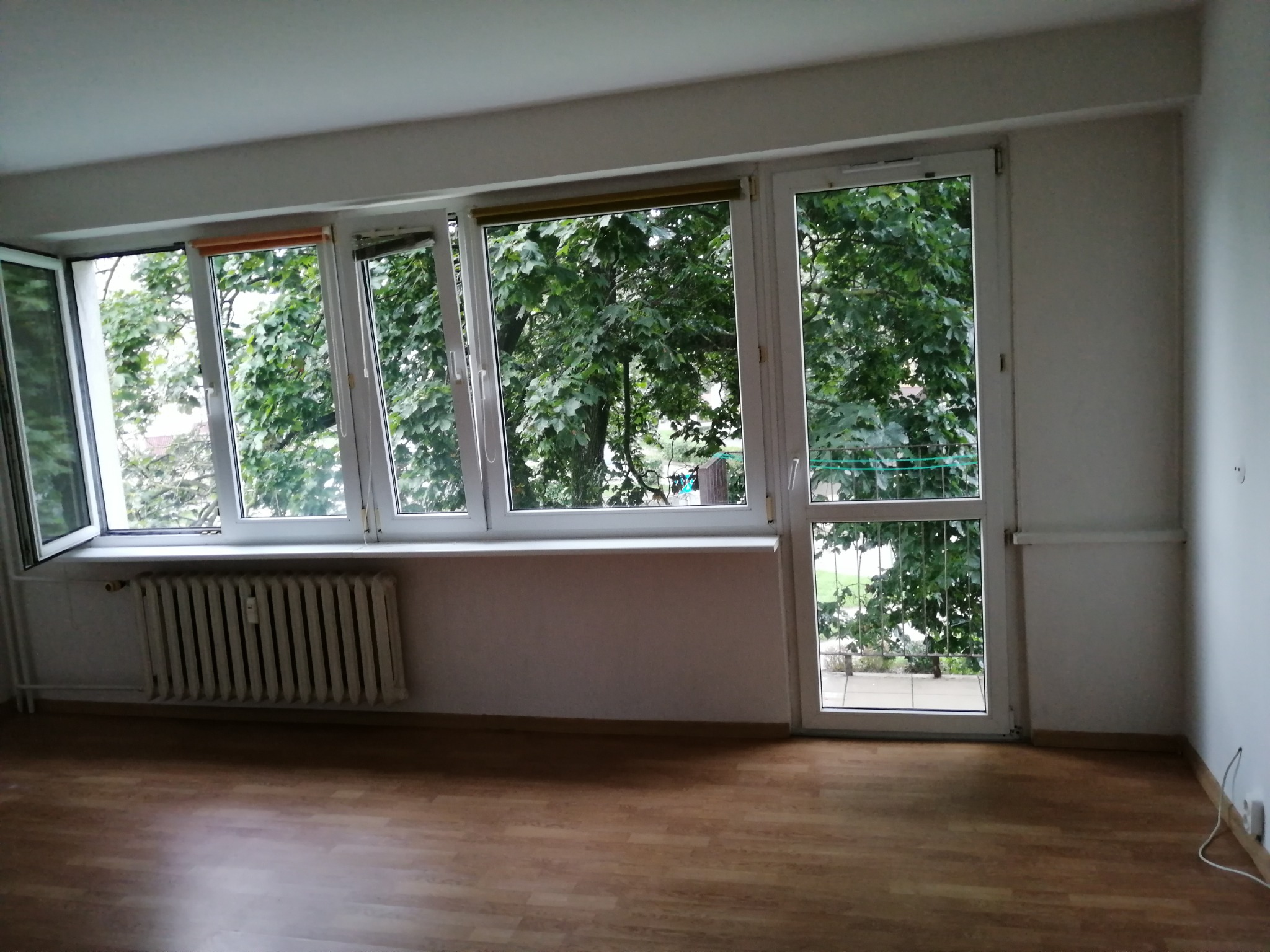 Pusty pokój z parkietem, grzejnikiem pod oknem i dużymi oknami z widokiem na zieleń, przygotowany do prac wykończeniowych.