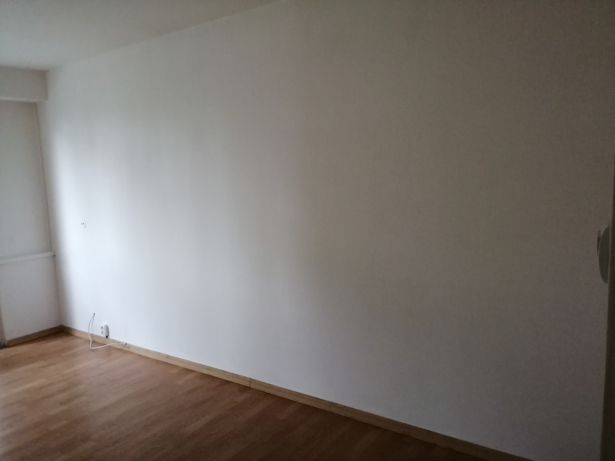 Pusty pokój z drewnianą podłogą, świeżo pomalowanymi białymi ścianami i widocznym gniazdkiem elektrycznym z kablami przy podłodze.