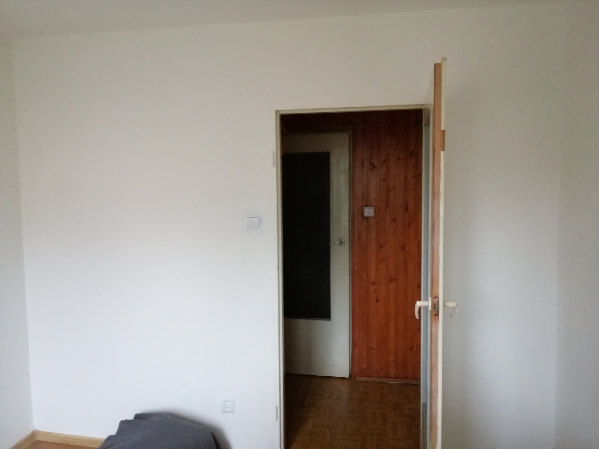 Pusty, jasny pokój z otwartymi drzwiami ukazującymi wnętrze z panelami ściennymi i ciemnymi drzwiami, widoczny fragment podłogi i ściany z gniazdkiem elektrycznym oraz włącznikiem światła.