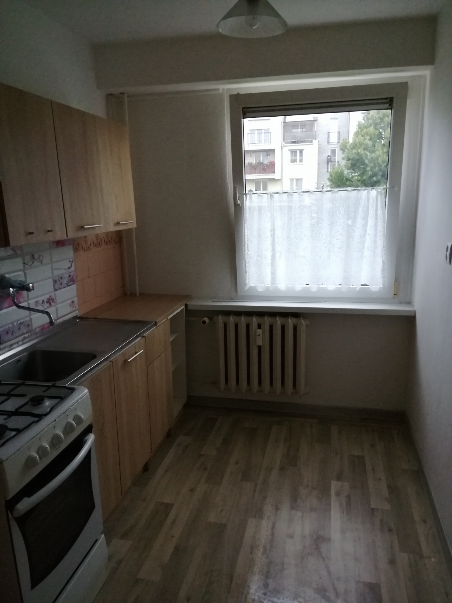 Wnętrze małej kuchni po remoncie: panele podłogowe imitujące drewno, jasne szafki z metalowymi uchwytami, okno z białą firanką, grzejnik pod oknem, kuchenka gazowa z piekarnikiem.