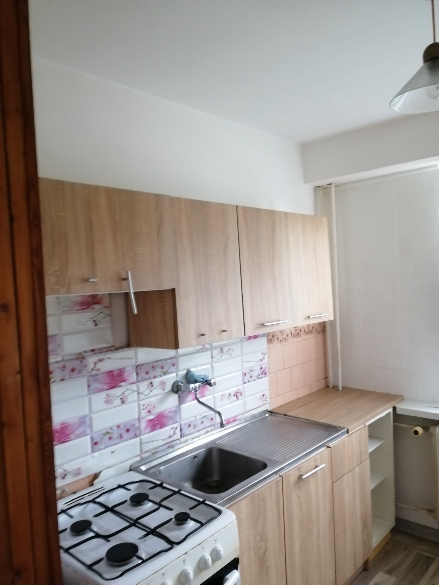 Kuchnia po remoncie: jasne, drewnopodobne szafki wiszące i stojące, płytki dekoracyjne z motywem kwiatowym nad blatem, biała kuchenka gazowa, stalowy zlew i grzejnik pod oknem.