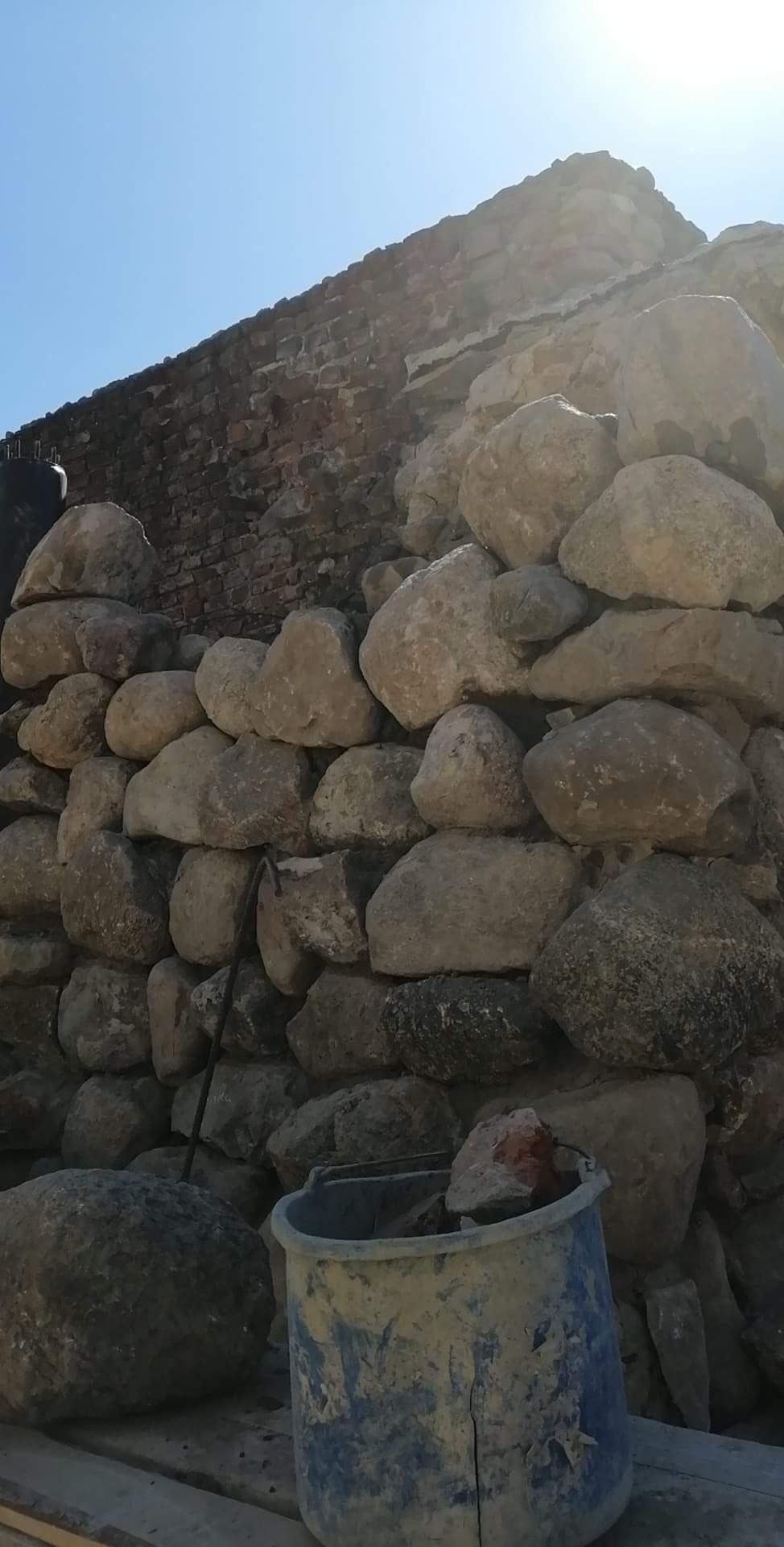 Wznoszenie ściany z kamienia polnego, widoczny fragment muru z cegły, niebieskie wiadro z zaprawą murarską na pierwszym planie, praca w toku.