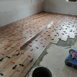 Układanie podłogi z płytek imitujących drewno, z użyciem poziomicy i czerwonych klinów dystansowych, widok na poddasze z oknem dachowym.