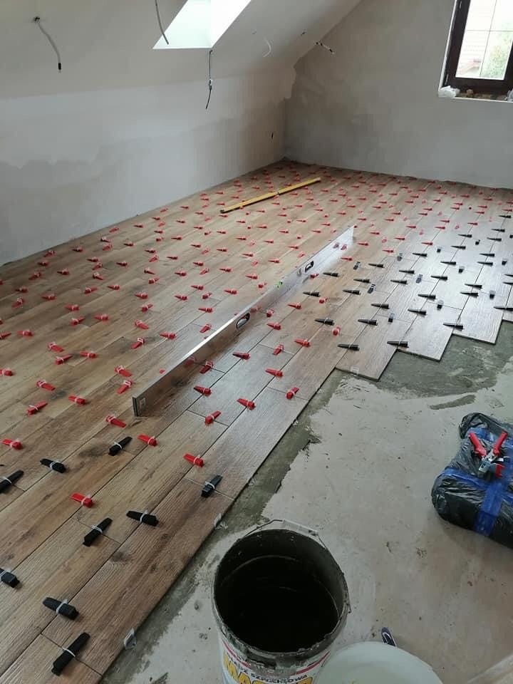 Układanie podłogi z płytek imitujących drewno, z użyciem poziomicy i czerwonych klinów dystansowych, widok na poddasze z oknem dachowym.
