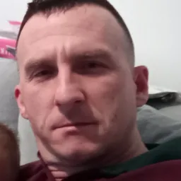 Portret mężczyzny z krótkimi, ciemnymi włosami, w bordowej koszuli, na tle jasnej ściany i fragmentu szarej kanapy.