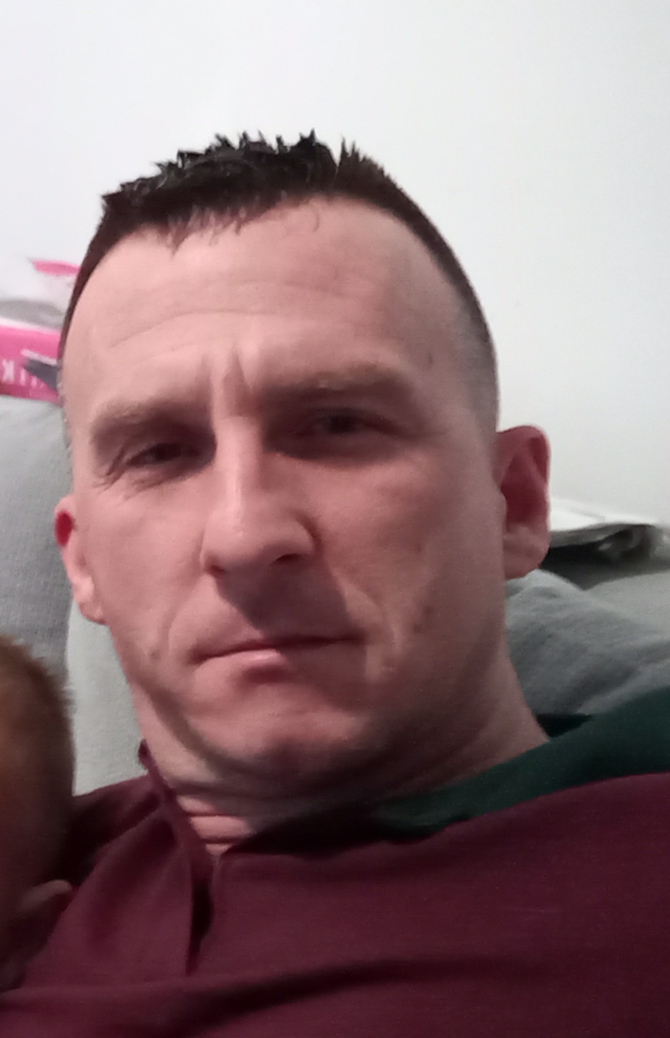 Portret mężczyzny z krótkimi, ciemnymi włosami, w bordowej koszuli, na tle jasnej ściany i fragmentu szarej kanapy.