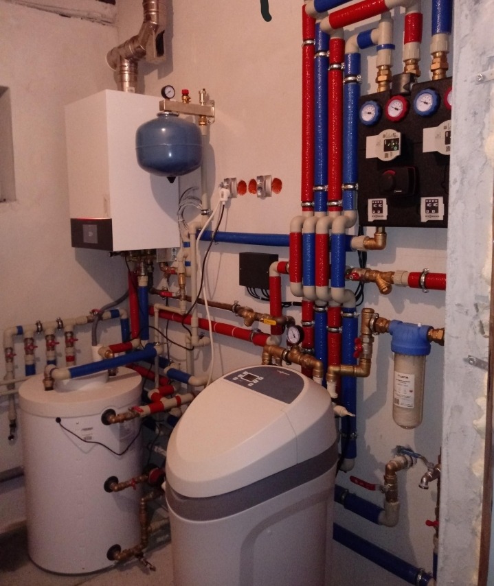 Kompleksowa instalacja hydrauliczna w kotłowni z piecem, zbiornikiem CWU, zmiękczaczem wody i filtrem narurowym, rury w izolacji czerwono-niebieskiej.