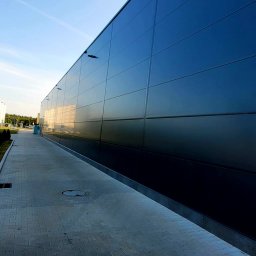 MARPAP - Ściana hali przemysłowej z paneli warstwowych, widok z dołu, z chodnikiem z kostki. Minimalistyczny design, odbicia nieba na panelach.