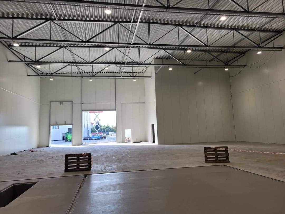 Wnętrze hali magazynowej z widoczną konstrukcją stalową dachu, ścianami z jasnych paneli, otwartymi bramami wjazdowymi i paletami na betonowej posadzce.