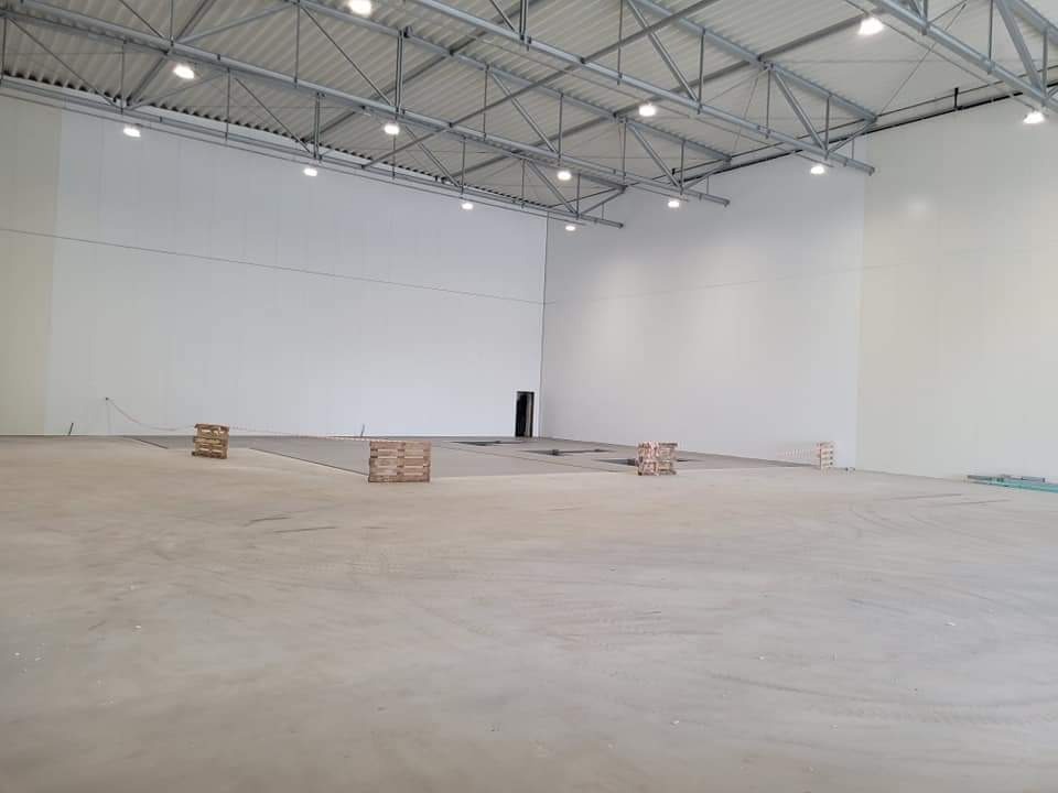 Wnętrze hali magazynowej z widoczną konstrukcją dachu, białymi ścianami z płyt warstwowych i betonową posadzką, na której leżą drewniane palety.