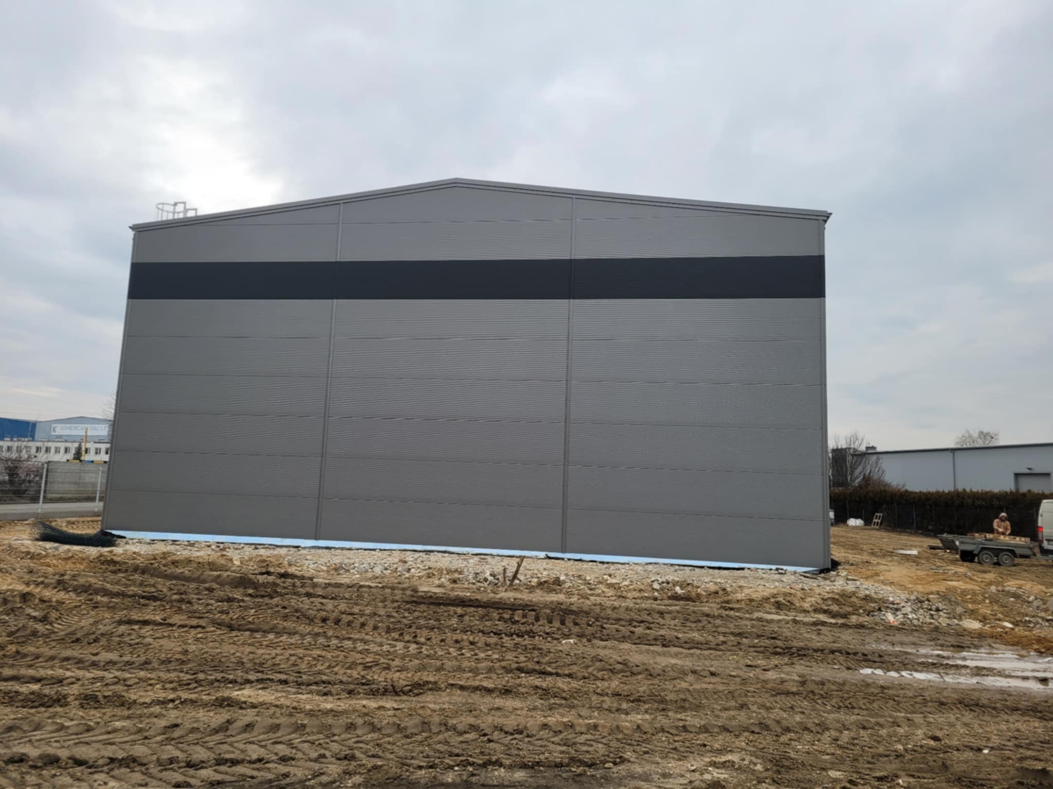 Nowo wybudowana hala magazynowa z szarej blachy trapezowej z ciemnym pasem na środku, widoczna na tle pochmurnego nieba i placu budowy z ziemią i gruzem.