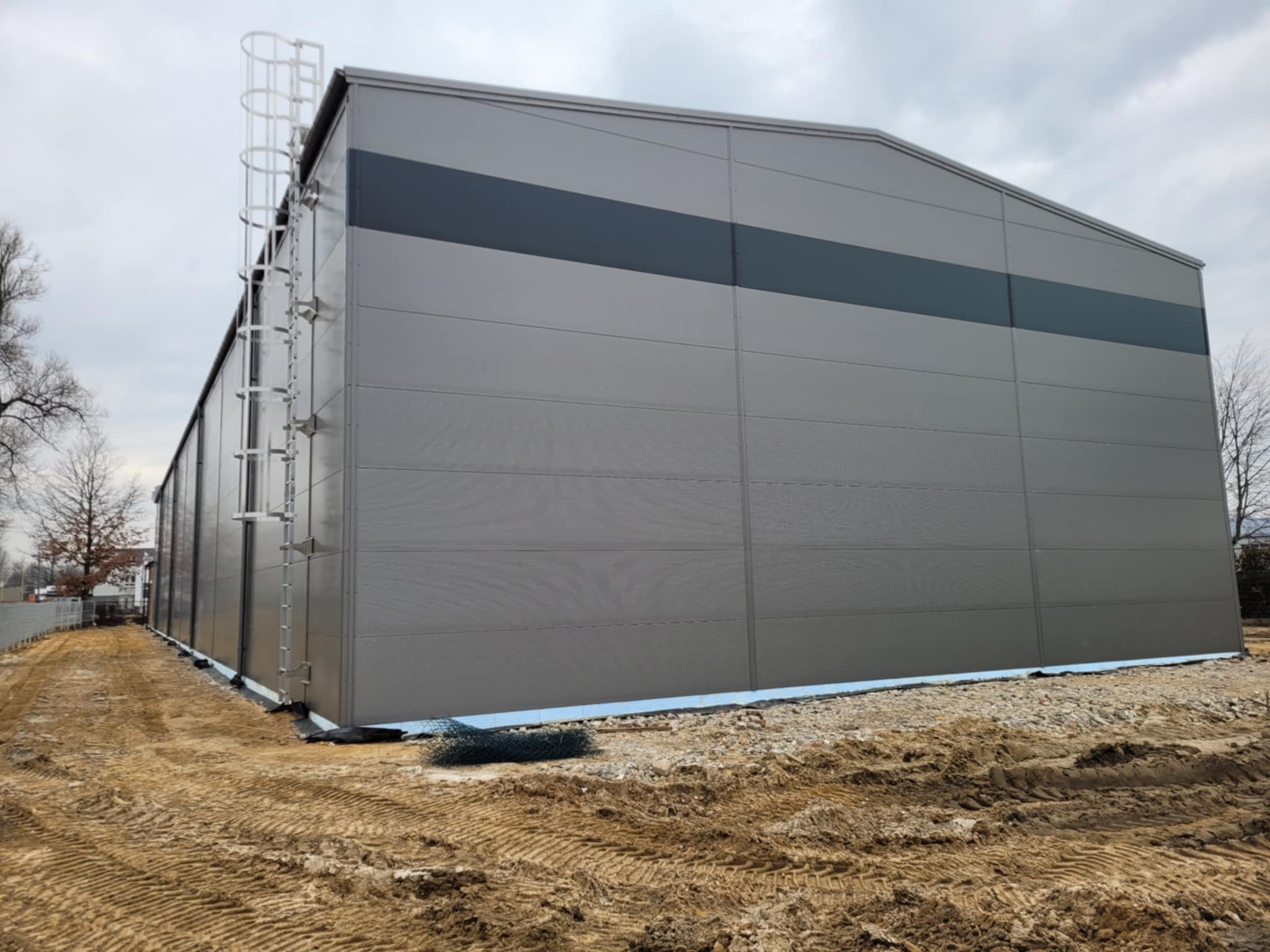 Szara hala przemysłowa z płyt warstwowych, widoczna drabina zewnętrzna, teren wokół w trakcie prac ziemnych.