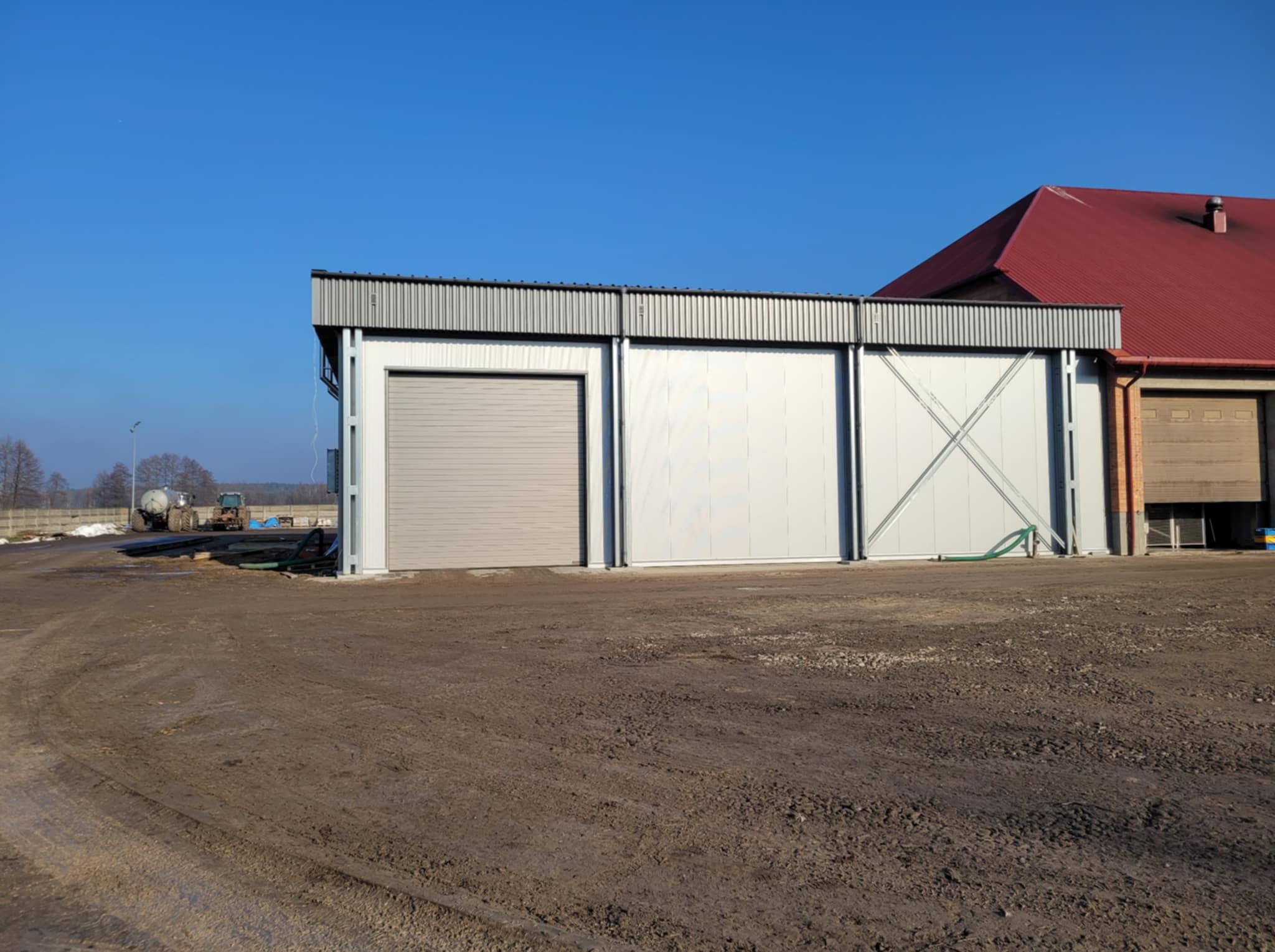 Gotowa hala z szarej płyty warstwowej z bramą rolowaną, widoczne elementy konstrukcji stalowej, obok budynek z czerwoną dachówką, na podłożu błoto i traktor w tle.